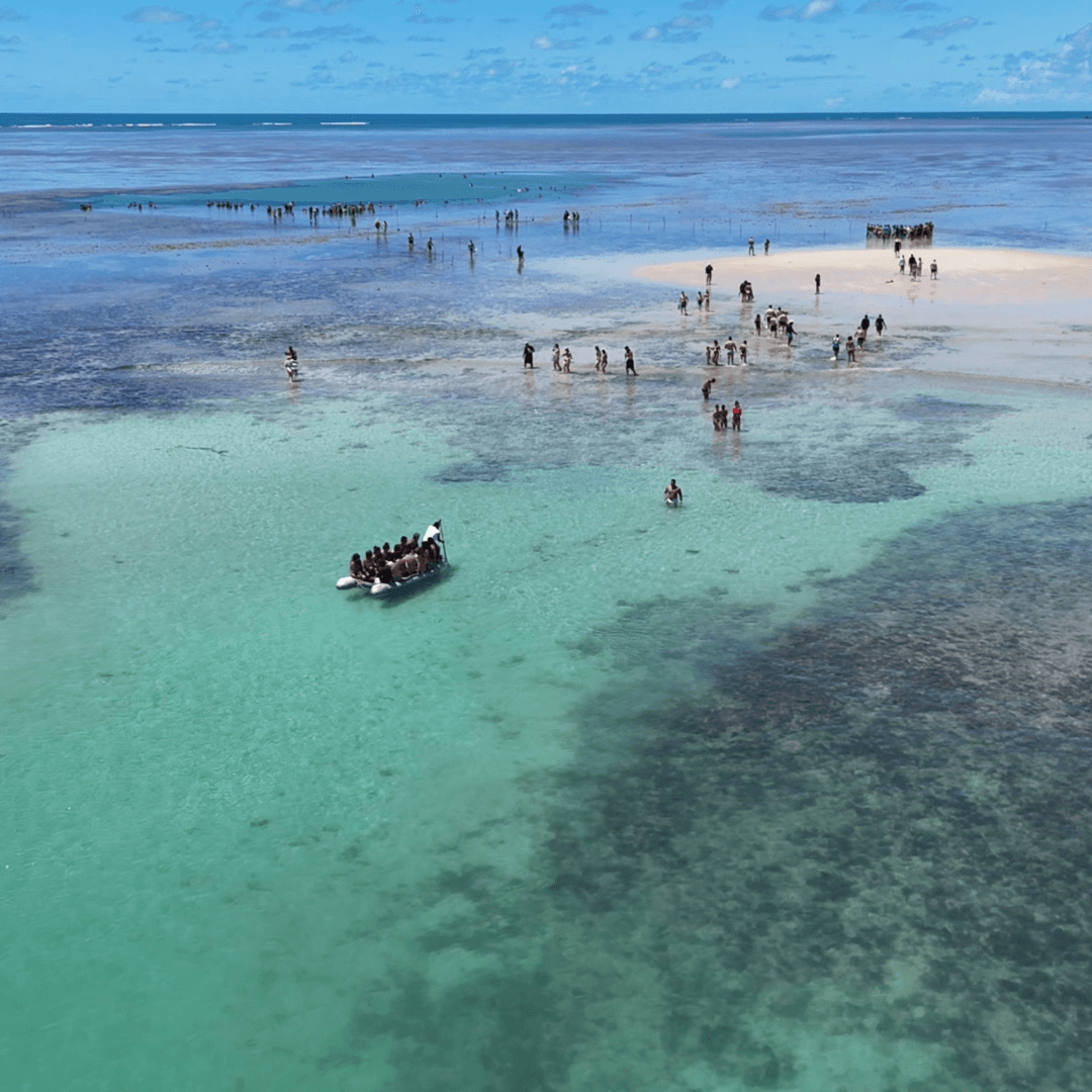 Vista aérea com barco inflável e grupos de pessoas em águas rasas.