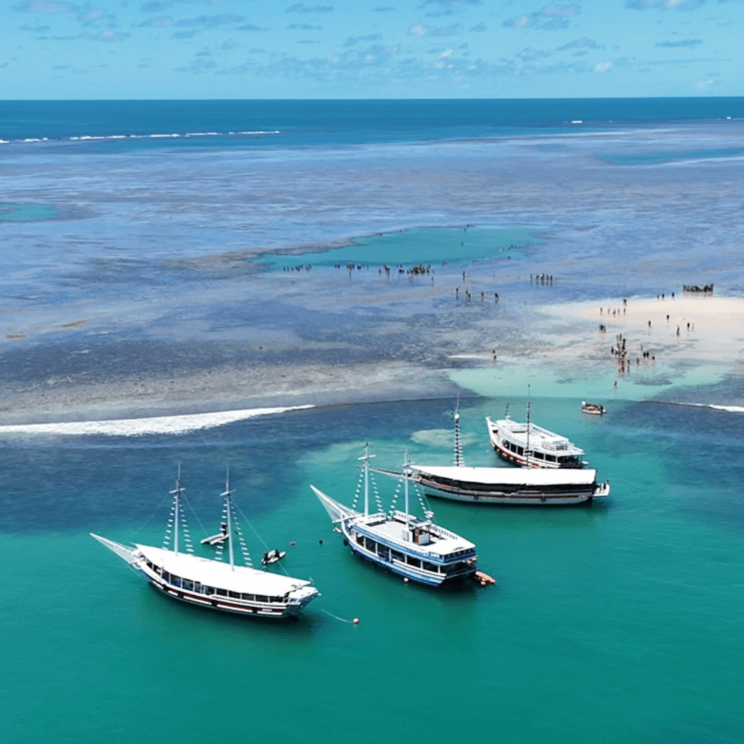 Vista aérea de águas turquesas com barcos ancorados e pessoas em piscinas naturais.
